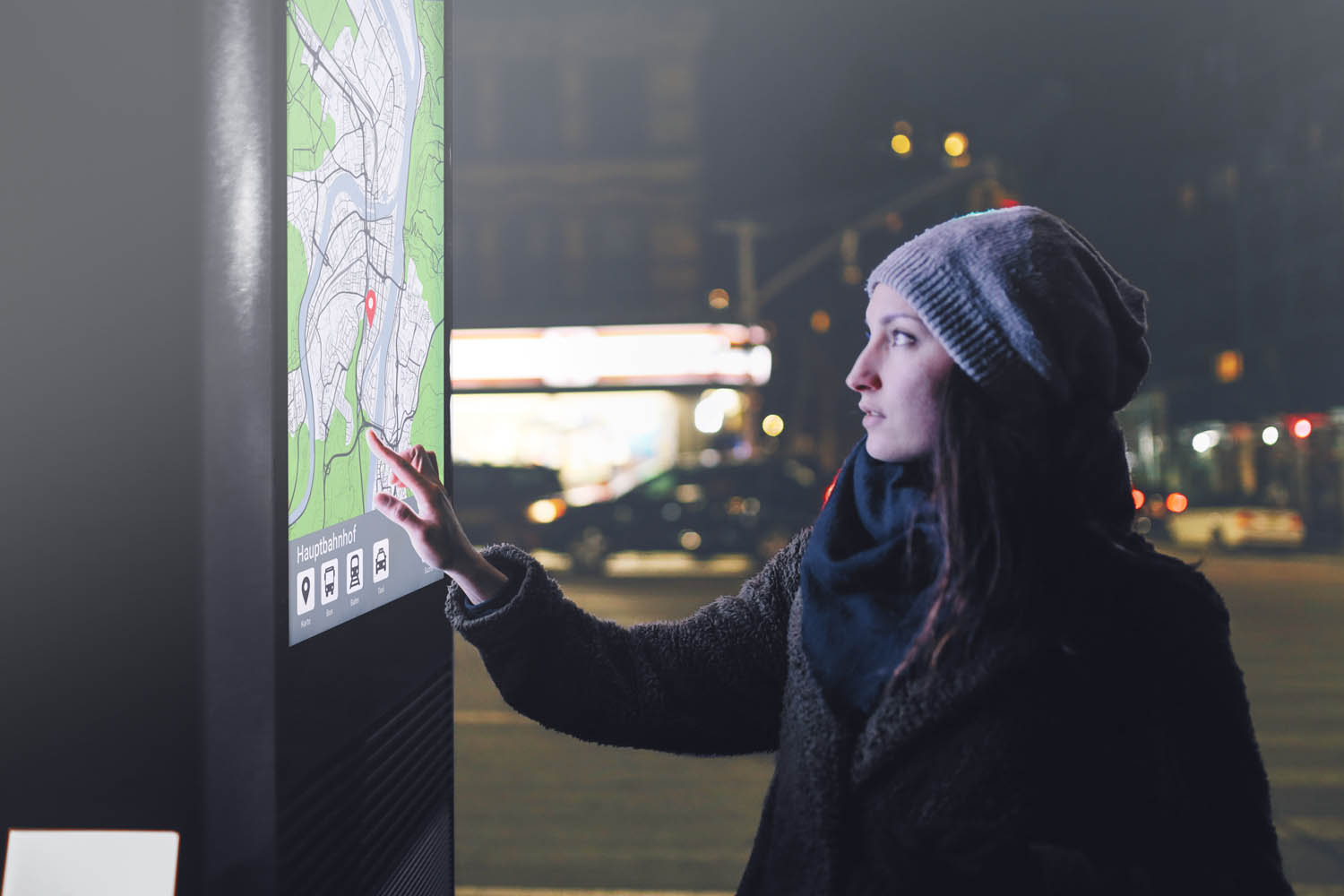 Eine Frau tippt, in der Stadt nachts, an einem Bahnhof, auf eine interaktive outdoor Stele welches eine Karte anzeigt