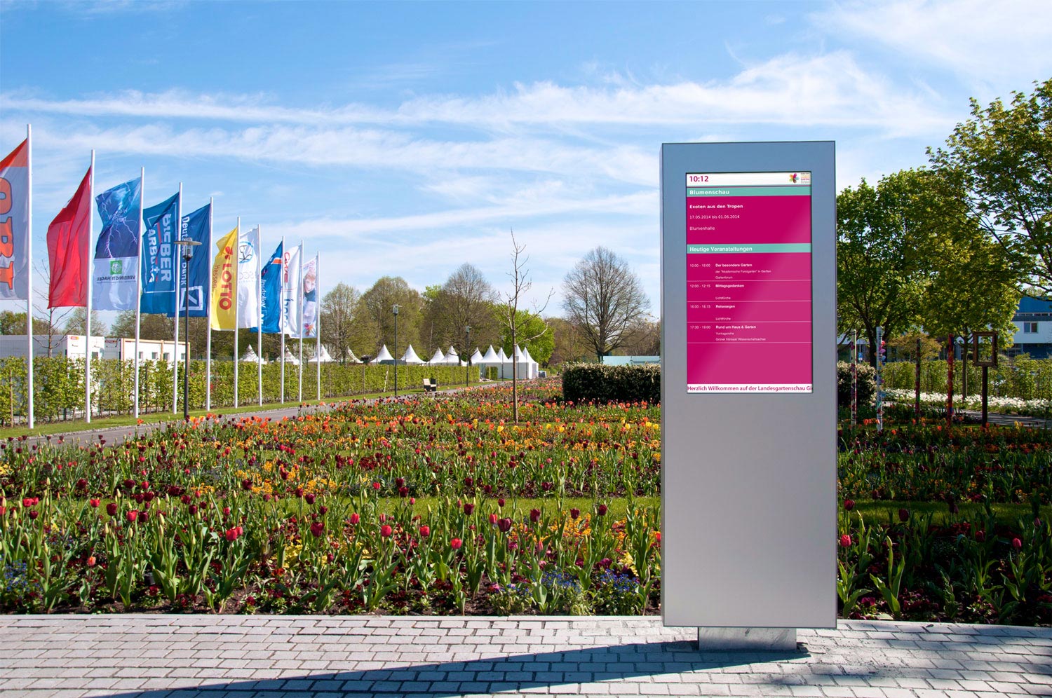 Eine Digitale Informationsstele, auf einem gepflasterten Bürgersteig, vor einem Blumenfeld mit blauen Himmel.